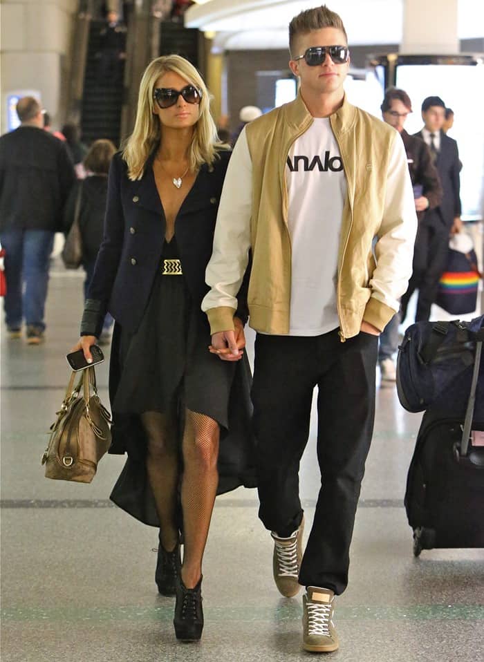 Paris Hilton looks effortlessly chic in a black high-low dress, fishnet stockings, and platform heels as she strolls through the airport hand-in-hand with River Viiperi