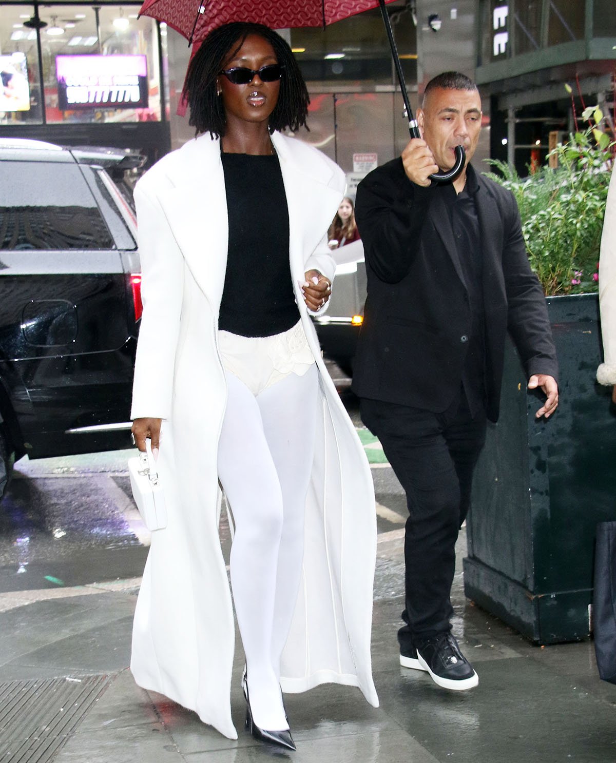 Jodie Turner-Smith wears an unconventional winter look, pairing a black knit sweater with a white coat, white opaque tights, and white diaper shorts