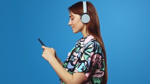 Side view of young student girl using mobile phone and looking at screen, wearing headphones while listening to music, isolated on blue background. People, lifestyle and emotion concept