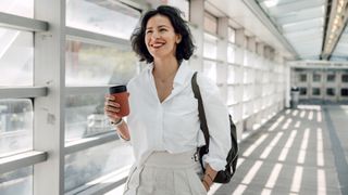 Woman walking with reuseable coffee cup down a hallway wearing trousers, a white shirt, and backpack