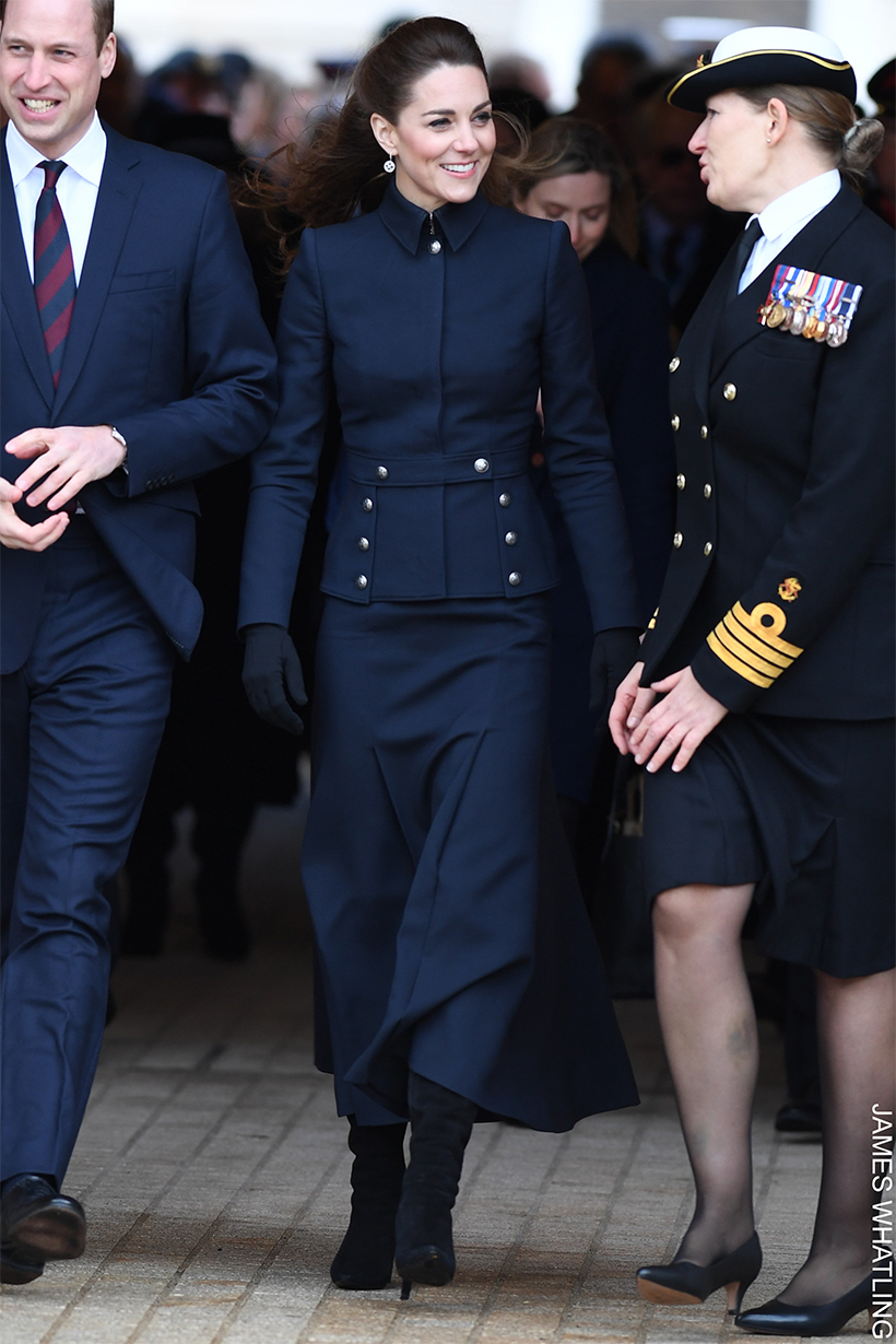 Kate Middleton wearing a navy military-style jacket with silver buttons, a matching midi skirt, and black heeled boots, walking outdoors alongside others.