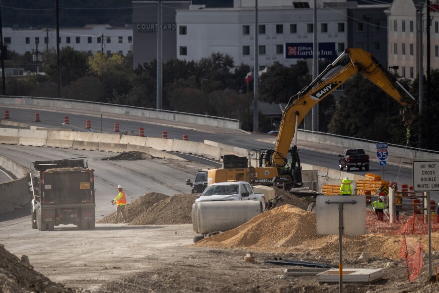 Construction on Interstate 35 as pictured from the Parmer Lane bridge in October. A person in high-visibility clothing is walking toward a dump truck. Pylon cones are in the background, blocking part of the frontage road. Other construction machinery and large piles of dirt are visible in the foreground. 