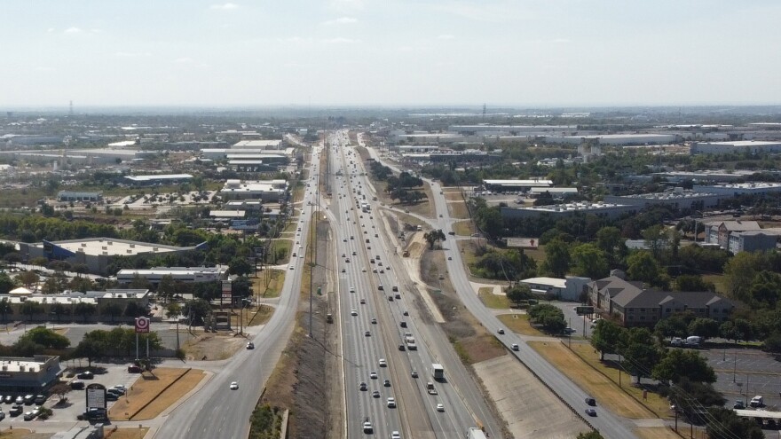 An aerial view of I-35 looking south from SH 45 North. Multiple lanes of traffic run in both directions, surrounded by commercial buildings, parking lots and scattered trees. 