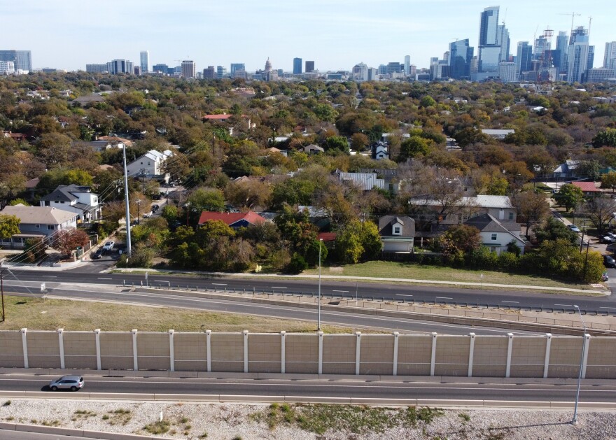 A view of a noise wall along MoPac looking eastward. The Clarksville neighborhood, seen as a mix of single-story homes and trees, is behind the noise barrier. Part of the downtown Austin skyline is visible in the distance. 