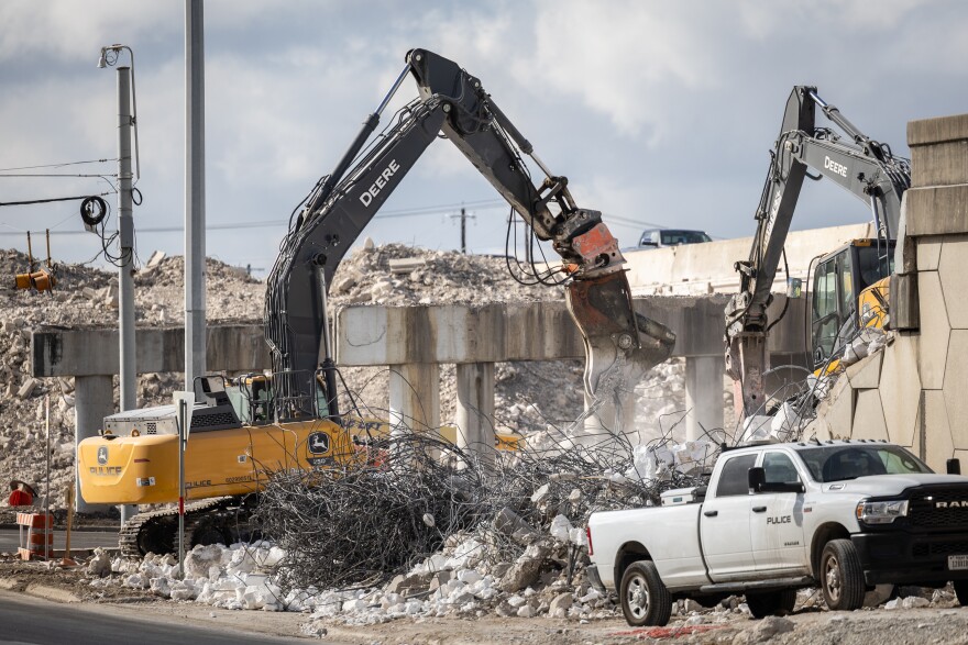 Demolition work at the I-35 and Howard Lane intersection showing large construction excavators breaking apart a concrete bridge with concrete debris and rebar visible. 