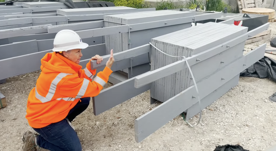 A woman in an orange high-visibility jacket and white hard hat crouches beside large gray bat boxes. She is holding and examining one of the boxes. Some construction materials are scattered around.