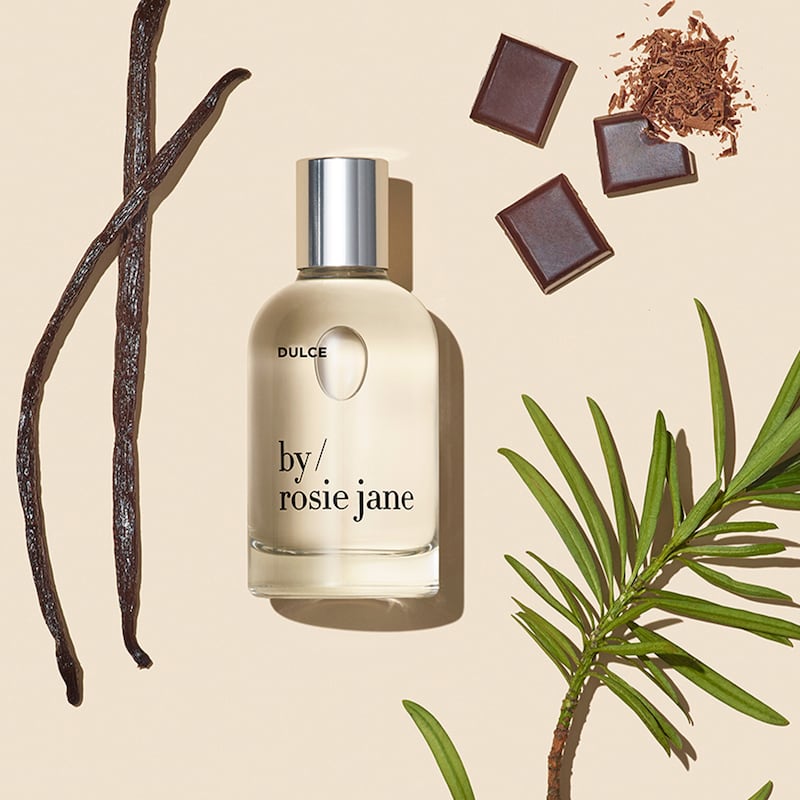 A clear bottle of fragrance, labeled Dulce by Rosie Jane, lays on a beige background among ingredients like chocolate and vanilla beans.