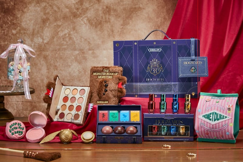 An array of Harry Potter-inspired beauty products by SheGlam, including a Book of Monsters eyeshadow palette and Golden Snitch highlighter, are displayed on a wooden table.