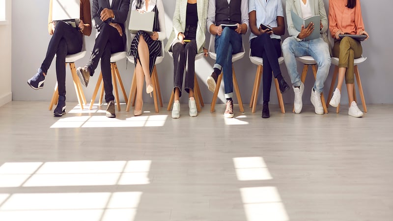 Diverse people waiting in line for appointment, job interview or work meeting. Group of male and female candidates sitting in corridor, reading, using different devices. Low section shot of human legs