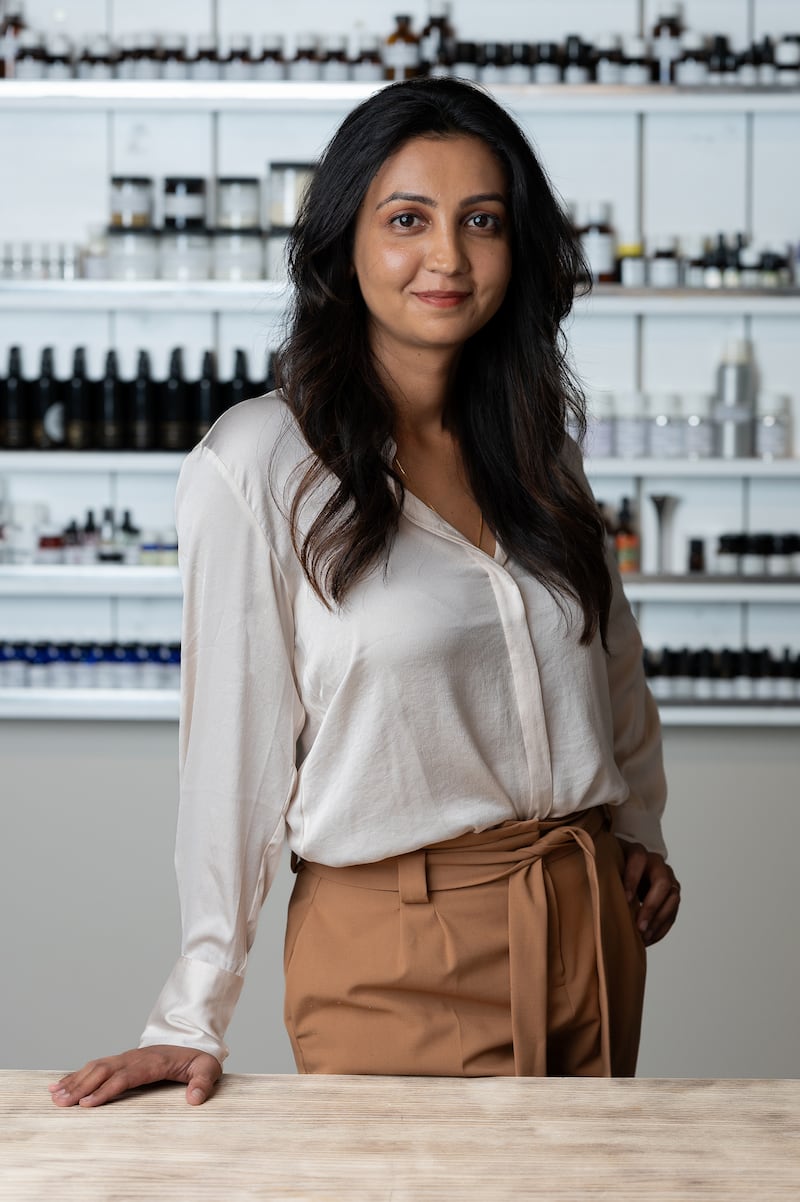 A woman Krupa Koestline, wearing a white buttondown blouse, brown pants with a tie belt, and her long dark hair over her shoulders, poses in front of shelves of cosmetic skincare samples.