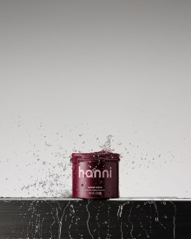 A small, squat burgundy aluminum jar of Hanni Splash Salve, an in-shower shaving aid, is splashed with water.