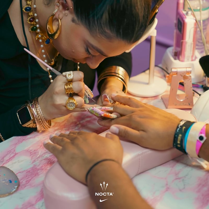 A woman wearing various wristbrands for different events is getting her nails done. The woman doing her nails wears lavishly long acrylics in Floridian sunset tones, some branded with the word NIKE or the brand's logo. She also wears a stack of gold bracelets and several chunky rings, one is a capital P.