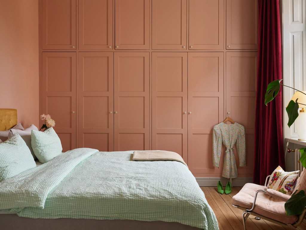 A monochromatic wall-to-wall wardrobe in terracotta stands out in a simple bedroom with red velvet curtains