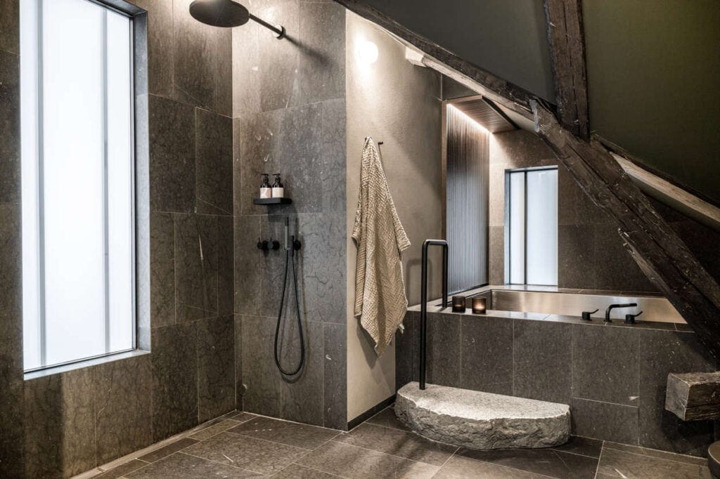 A dark and earthy bathroom with grey limestone tiles, dark ceilings, and black fixtures