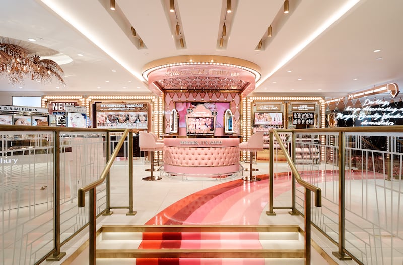 The first floor of Charlotte Tilbury's new Covent Garden store. At the center is a pink half moon-shaped counter with pink bar seats. Vanity-lit makeup displays are on either side, and a pink-hued rainbow leads from the front door to a mirrored lounge.